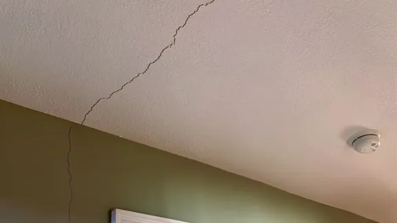 A fine hairline crack running across a lightly textured painted ceiling toward the corner where the ceiling meets the wall, with a round white smoke detector mounted nearby
