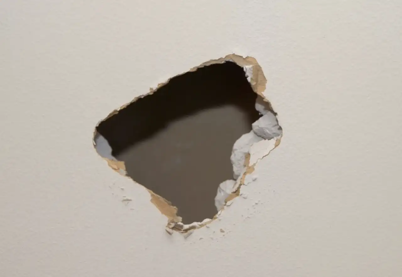 Jagged impact hole broken through a painted interior drywall wall, torn paper facing and crumbling gypsum around the opening exposing the dark cavity behind
