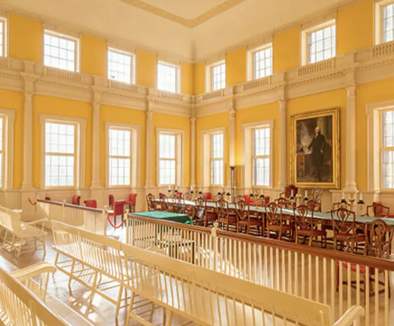 Historic Connecticut state government chamber with tall windows, columned plaster-finished walls, a portrait, and a spectator gallery above rows of desks