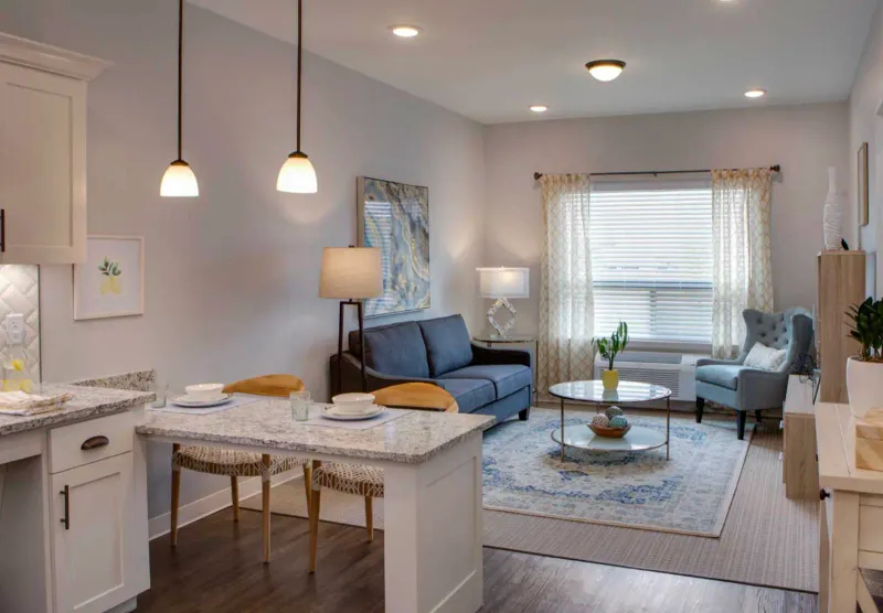 Senior-living apartment unit with finished painted drywall, recessed and pendant lighting, an open kitchenette and dining table beside a living area with a sofa, armchair, and curtained window